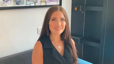 Dr Rachael Molitor in a sleeveless black top sat on a grey sofa. She has long brown hair. On a wall behind her is the bottom of some arwork - the words Foleshill Road can be seen.