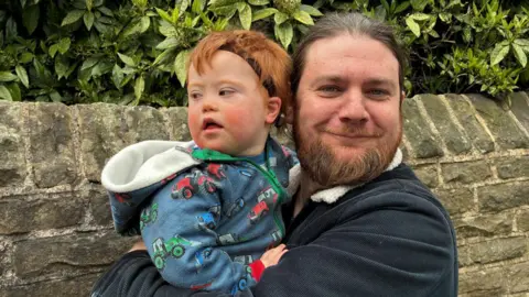 A man with brown hair and a beard stands with his back to a stone wall with bushes behind it. He is looking towards the camera and smiling and is holding a toddler in his arms. The child is wearing a hooded jacket in blue. 