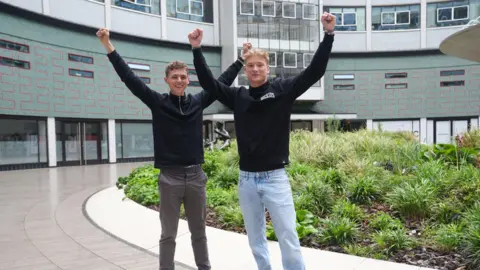 BBC Alfie Watts and Owen Wood standing outside in front of an office building. They both have their hands held above their heads and are wearing dark tops, jeans, and trainers and are smiling.