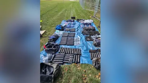 Dozens of packets of drugs laid out on a blue groundsheet in a field.
