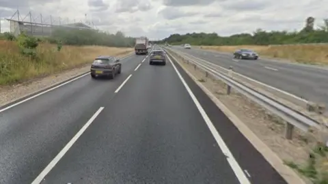 A view from a Google vehicle on the A12 dual carriageway, seen from the right hand lane of the left hand carriageway. It is lined with grass verges. A few vehicles are moving on the road and there is a stadium to the left.