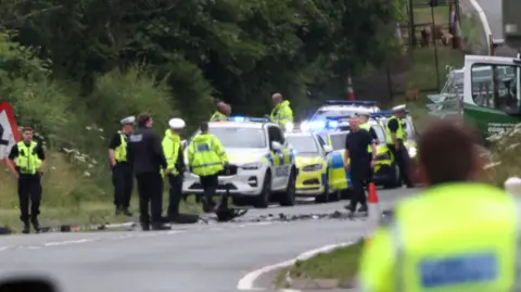 The scene of a crash on the A1 near Causey Park. A mass of police officers are standing in the road, which has been cordoned off, with police cars parked with their blue flashing lights on. Vehicular debris is strewn across the road.