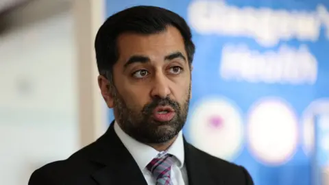 PA Media A man with a dark hair and beard speaks in front of a blue sign. He is wearing a black suit, white shirt and purple tartan tie.