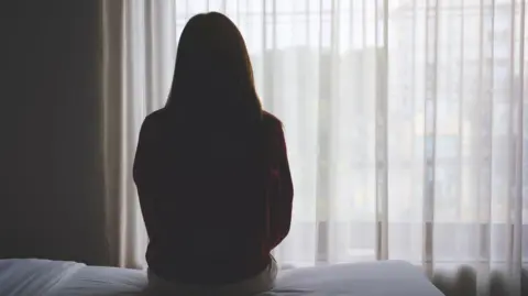 Getty Images The back of a woman sitting on a bed looking out a window with curtains on it. 