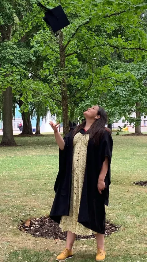 Abbie Tutt  Abbie Tutt in clothing worn for graduation, throwing her hat in the air in front of a park with a tree directly behind her.