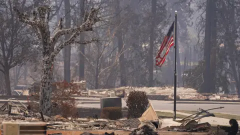 A burned United States flag hangs from a charred flagpole - burned trees and remnants of destroyed properties can be seen on either sides of the road and the air is hazy 