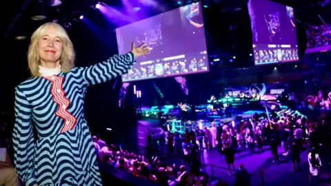 City Hall Justine Simons wearing a blue and white striped dress, smiles and gestures towards large screens showing a video game, above a crowded esports arena lit in blue and purple.