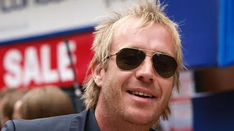 Rhys Ifans wearing sunglasses and smiling. He has blonde hair and a grey suit