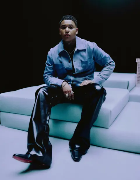 Kamal Shaquille Promotional photo of KWN, sitting down in a denim shirt and black leather trousers