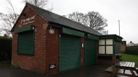 The cafe in Paddy Freeman's Park, Newcastle. It is a small, red brick unit, with dark green shutters. Above one of the shutters, at the end of the building, there is white signage that contains a duck and the words The Park Cafe @ Paddy Freeman's. There is a wooden picnic bench out front and some leafless trees in the background,