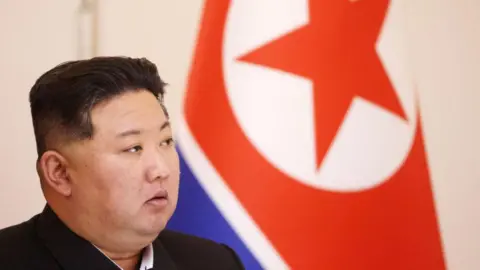 Kim Jong Un looking to the side. Behind him is a large North Korea flag.