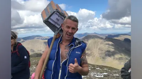 Nigel Howell A man with grey hair in a blue unzipped gilet giving a thumbs up with his left hand and in his right holding a wooden pole and a carrier full of bricks over his shoulder, with a sign on saying Justgiving The Hodfather. He's standing on the top of a mountain with a range in the background. There is some snow on the ground and the sky is blue with some clouds.
