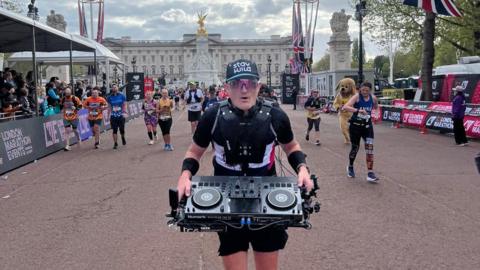 Gus Fraser runs towards the camera with his DJ kit attached to the harness and behind him is Buckingham Place and other London Marathon runners