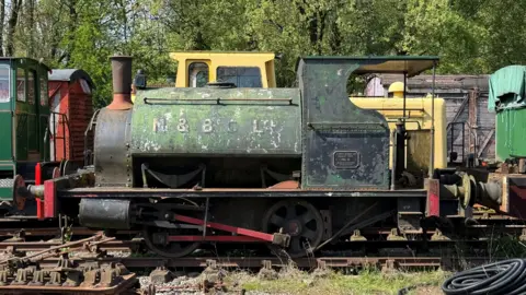 A shabby-looking locomotive with paint flaking away. It is green in colour with white lettering just visible. It is sat on rails among a number of other carriages and locomotives.