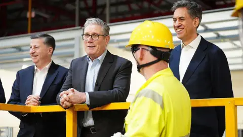 Getty Images Two people in business attire stand on an elevated platform inside an industrial facility, speaking with a worker wearing a high-visibility jacket and yellow safety helmet in the foreground. Yellow safety railings and factory equipment are visible in the background.