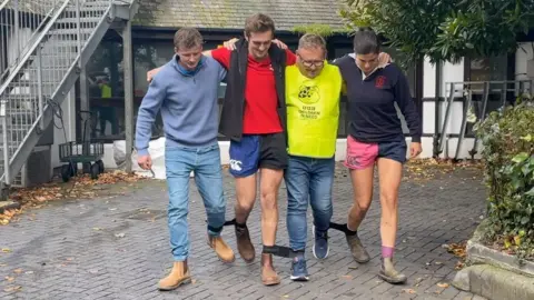 Three men and one woman (right), with their legs tied together with black elastic so they are three-legged. They have their arms around each other.