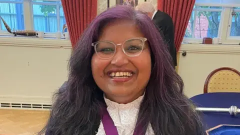 Facebook A woman with faint purple hair is smiling enthusiastically at the camera. She is inside what appears to be a council chamber.