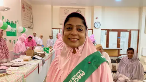 A woman in a pink-coloured Rida headdress with a green Macmillan Cancer Support sash across her shoulder.