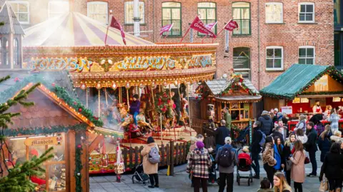 A carousel and market stalls in Leeds for the Christmas market. Many people wearing warm coats stand by the stalls watching people on the carousel