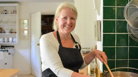 Jane Steward A woman smiles at the camera as she stirs a pot of food on a hob. She has blonde hair that has been tied back. She is wearing a black apron with a white long-sleeved top underneath. 
