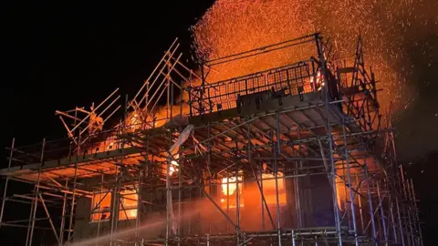 PVFB A house under construction surrounded by scaffolding is well alight and red flames and smoke and embers can be seen enveloping it and glowing against a night sky.