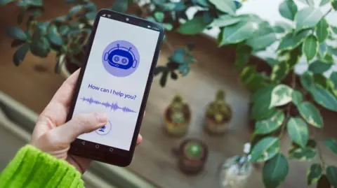 A person wearing a green jumper is holding a phone. On the screen is a purple logo showing a robot with a headset on. Underneath, it reads "How can I help you?". In the background of the photo is a table and some green plants