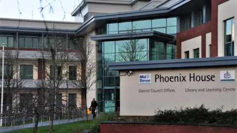 Lewis Clarke / Geograph The photograph shows the outside of Phoenix House, Tiverton. In the foreground is a sign that reads "Phoenix House, District Council offices". The building behind the sign has a glass entrance and the building appears to be three to four storeys high.