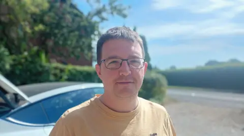 John Robinson, wearing glasses and a yellow t-shirt, speaks to the BBC as he loads his car
