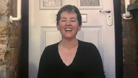 A woman with short hair in front of a white front door. She has small hoop earrings and a cross pendant on, and is grinning broadly. She is wearing a black top.