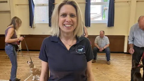 Pippa Apps is standing in a village hall with a group of dog owners and their pets behind her on leads.