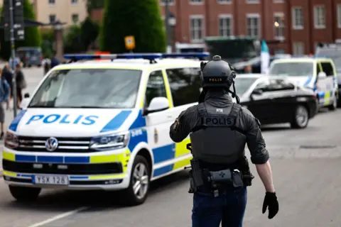 TT A police officer in tactical gear stands in front of a Swedish police car