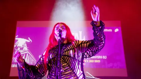 A woman with long red hair on stage in a spider's web outfit. There is a large screen behind her. 