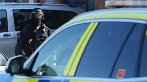 Eddie Mitchell A firearms officer on the scene at Woodside Road, Worthing. The officer is facing the camera and is wearing a police cap and ear defenders. His face has been pixellated to hide his identity.