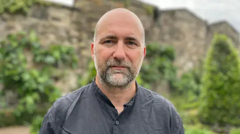 Simon Francis from the End Fuel Poverty Coalition is picture with a blank expression standing in a garden wearing a great shirt 