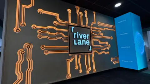 Ben Schofield/BBC A black wall with the Riverlane logo in the centre. The logo places the word 'river' over the word 'lane', both in lower case - apart from the 'l' in 'lane' - in a pale blue colour. In the top left corner of the logo is a right-angled lower case 'r' and in the bottom right corner is a capital 'l'. The logo is inside a black square. Orange and black lines, looking like tracks on a printed circuit board, emerge from behind the square, each ending in black circles. Next to the wall is a blue tower, on which is listed the time in Seattle, Boston, London and Singapore.