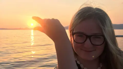 Family handout Lucy Curtis holds her fingers up to look as if she is pinching the sun as it sets while standing next to a lake. She has blonde hair and glasses.