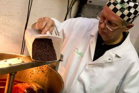 Duffy's Chocolate A man wearing a white lab-style coat and chef's hat tips cocoa beans into a large mixing bowl. 