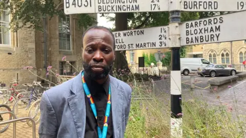 A man is standing next to a road sign. He is wearing a blue jacket and dark shirt. He looks concerned.