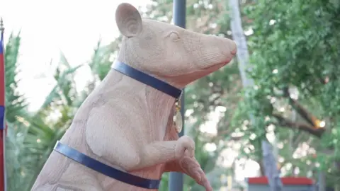 A statue of a giant rat with a black harness and medal dangling from its neck, made from stone, is pictured with trees in the background. 