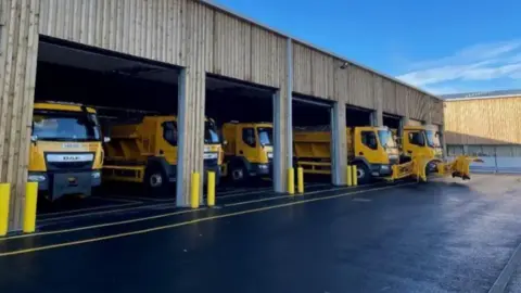 Wiltshire Council A view of Warminster salt depot with five yellow gritting vehicles parked at a holding station
