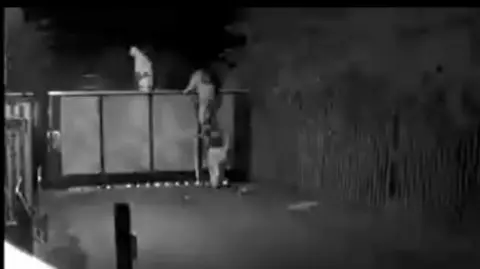 CPS A grainy black-and-white still from CCTV. Three people in white forensic-style suits are climbing over a gate. One is standing behind the gate, raised up on an unseen ladder looking down, and the other two are descending a ladder on the near side of the gate with their back to the camera.