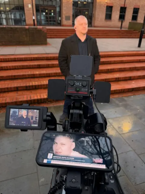 Rafal Zalewski A man in front of a camera on the steps of a crown court. A woman's face can be seen on a phone screen in the foreground.