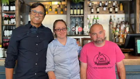 Rogerio is on the left, Amy is in the middle, and James is on the right. They're all looking and smiling at the camera. Behind them is the bar. 