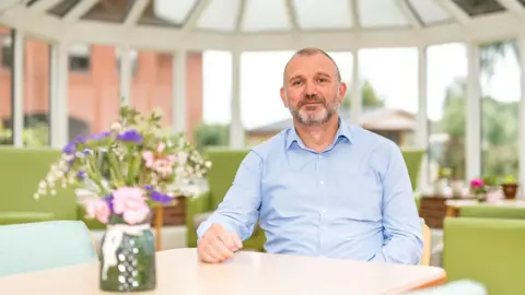 Birmingham Hospice CEO Simon Fuller sitting at a table in a conservatory at the hospice with a vase of flowers in front of him