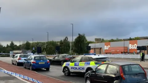 A road which looks like a dual carriageway with several cars and a police car, with police tape in front. There is a large building which says 'B & Q' in the background.