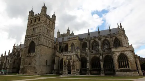 An outside shot of Exeter Cathedral. It shows all the architecture and the cathedral green. There is the main building and the tower which is at the heart of it. The weather is cloudy with a blue sky behind them. 