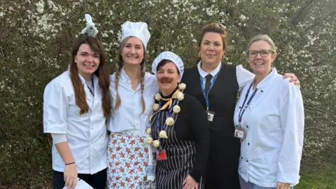 Abbeymead Five female teachers, all wearing chef whites and standing in a line in front of a hedge, smile at the camera