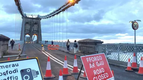 Menai Bridge with a 'road closed' sign and traffic bollards in the road, as some pedestrians cross over