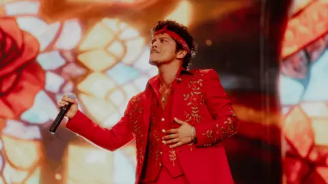 Bruno Mars holds a microphone and performs at Wembley stadium wearing a red suit with gold flower detail. He also wears a red headband.