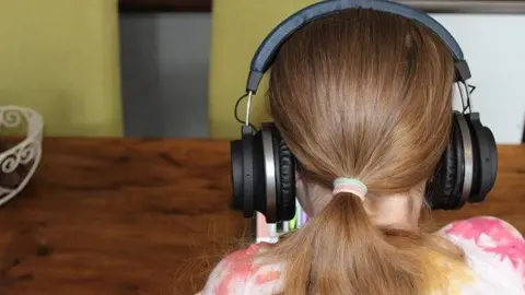 Link To Change A child, with its back to the camera, wearing headphones. They are by a wooden table, with long hair tied back.